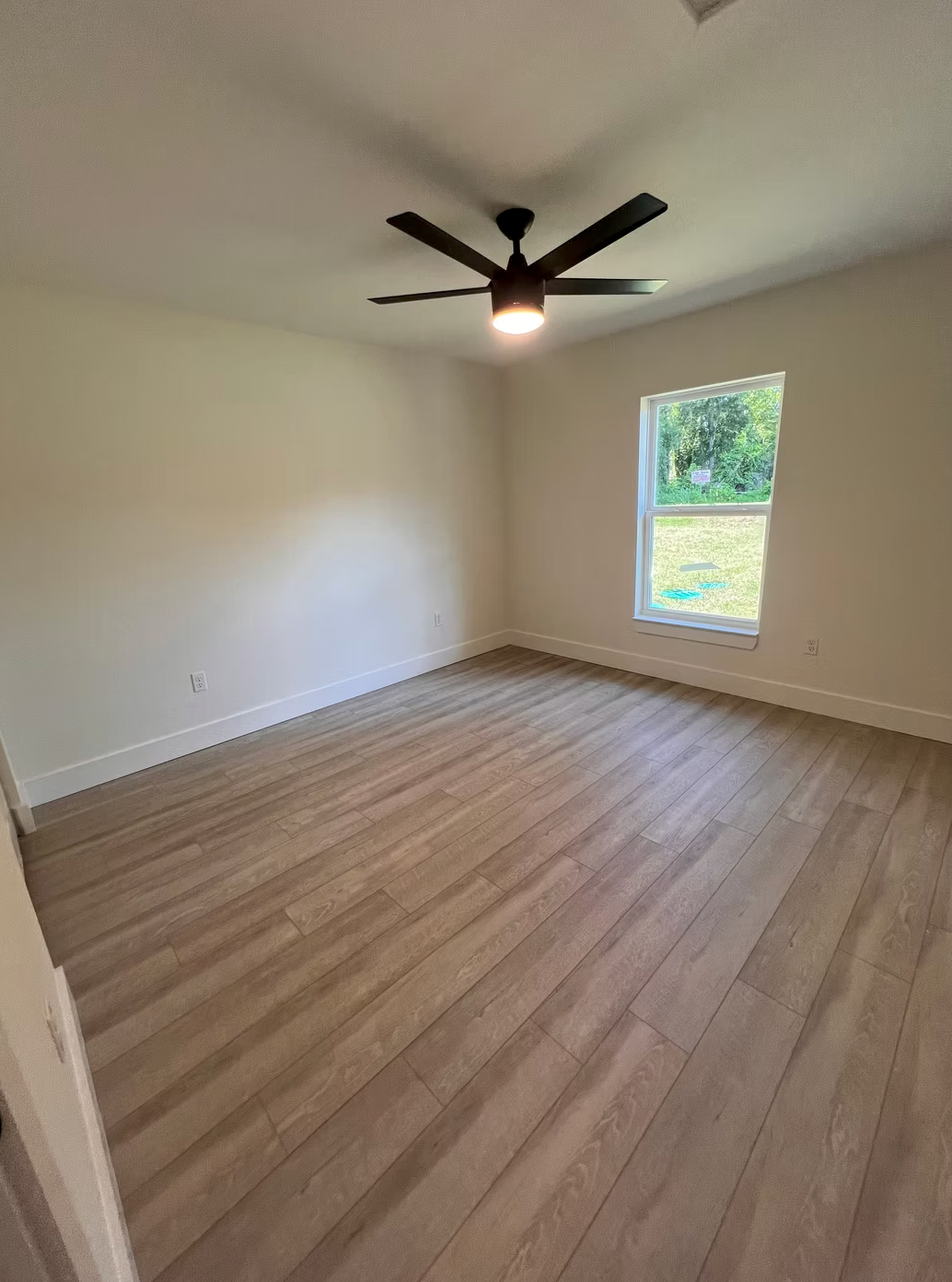 Empty bedroom with wood floors, white walls, ceiling fan, and a window letting in daylight