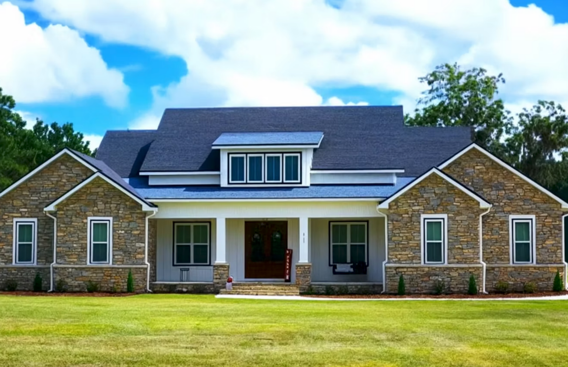 Large stone house with a dark roof, white porch, and manicured green lawn under a blue sky