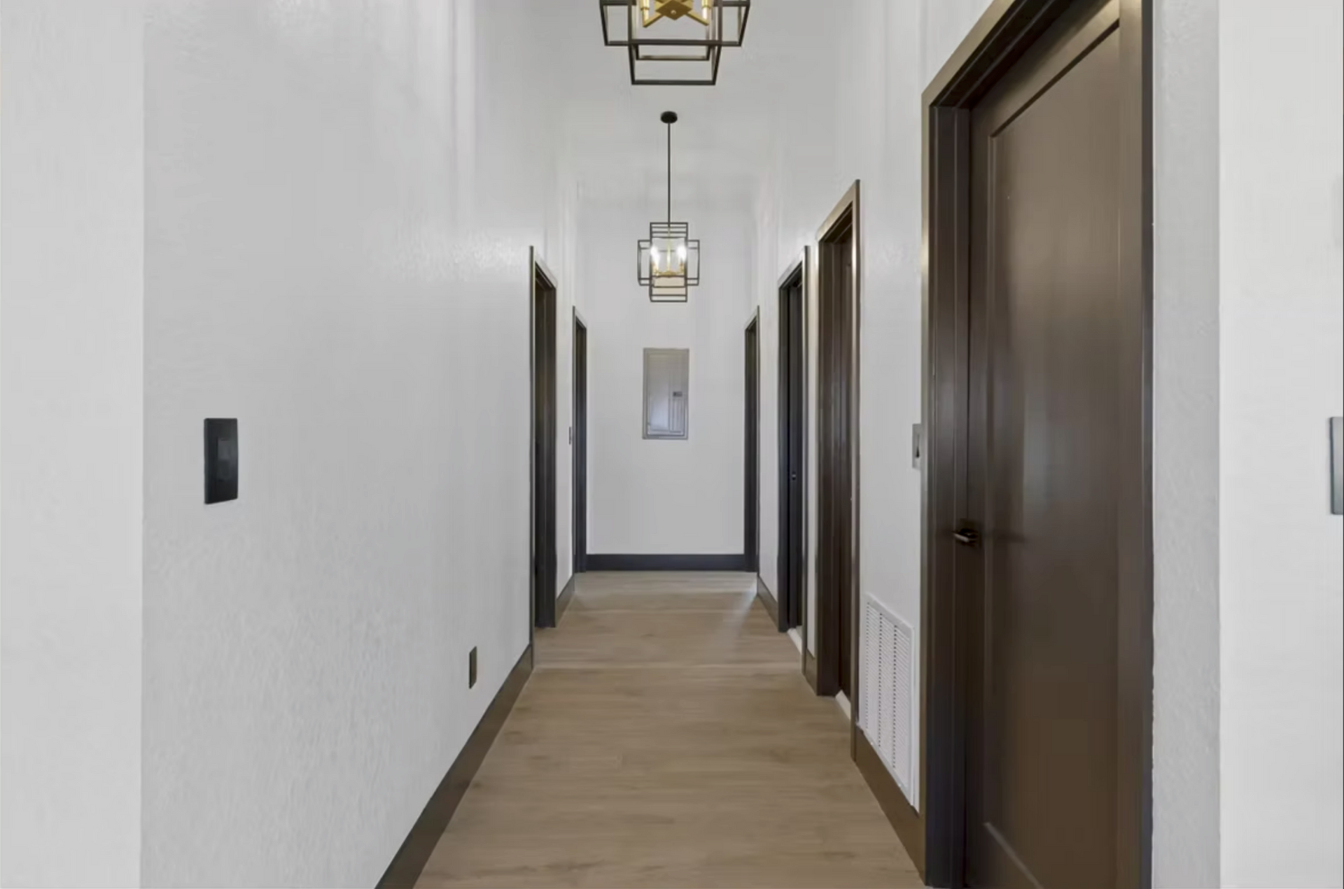 Long modern hallway with white walls, wood floors, dark doors, and hanging ceiling lights