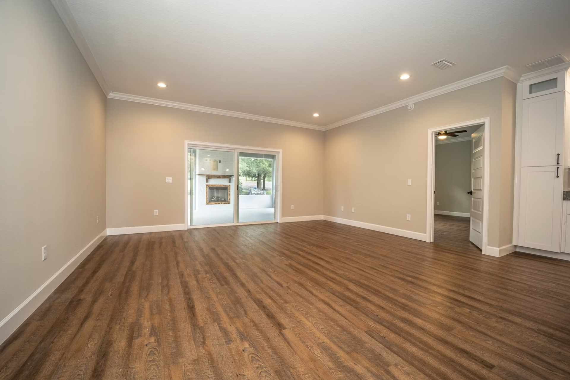 Empty beige living room with wood flooring, recessed lights, and sliding glass doors.