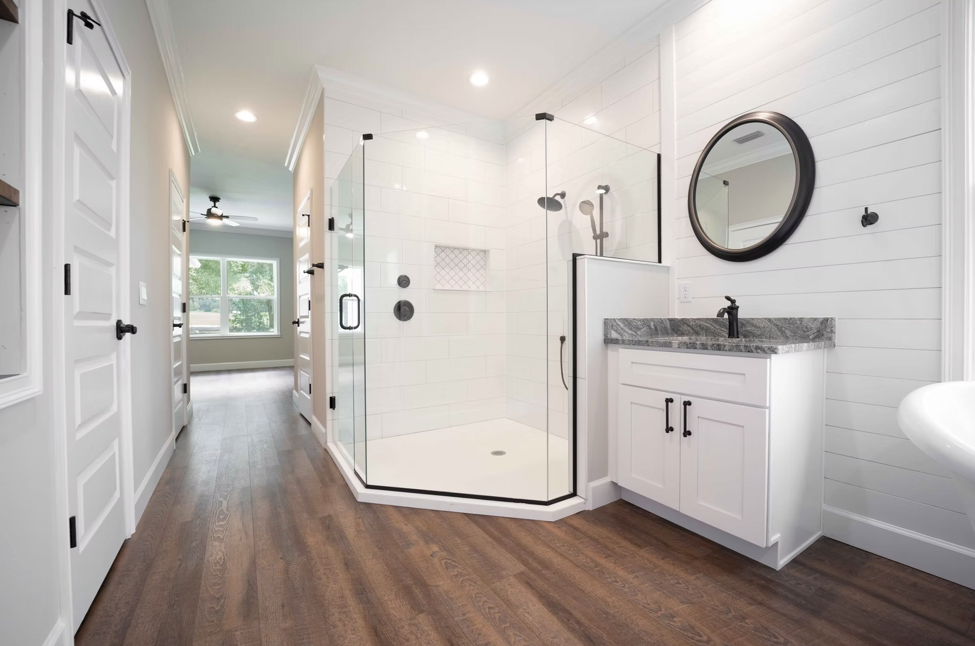 Bright bathroom with white walk-in shower, dark wood floors, and a vanity with round mirror.