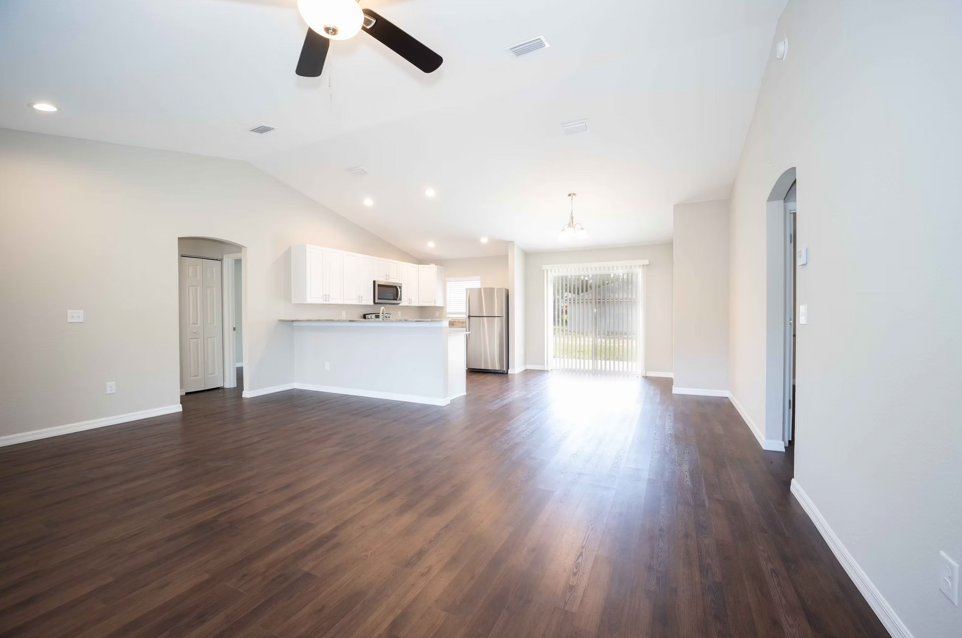 Spacious empty living room with dark wood floors, white walls, kitchen island, and bright sliding glass doors.