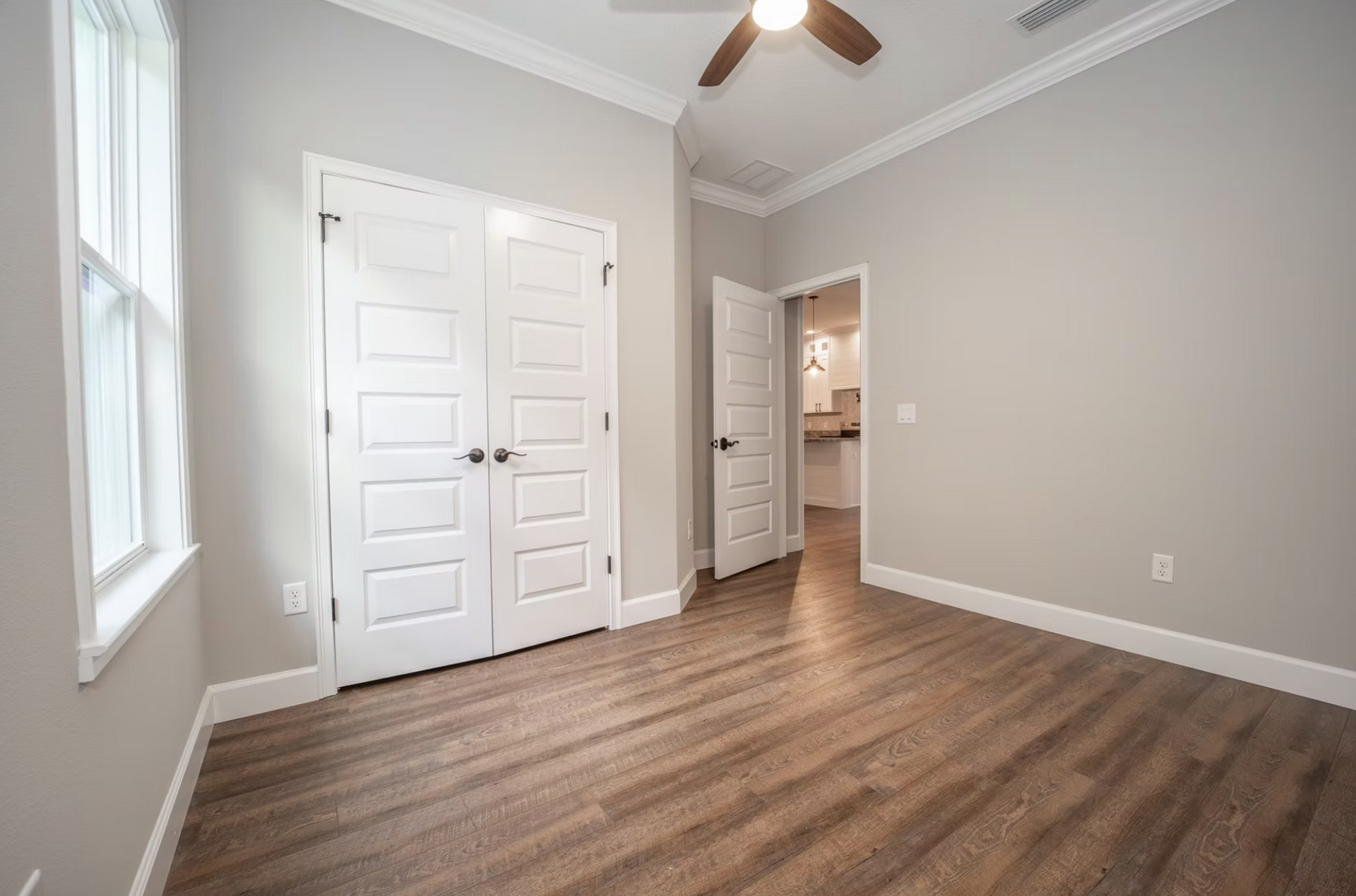 Empty gray bedroom with wood floors, white closet doors, ceiling fan, and an open doorway to a bathroom.