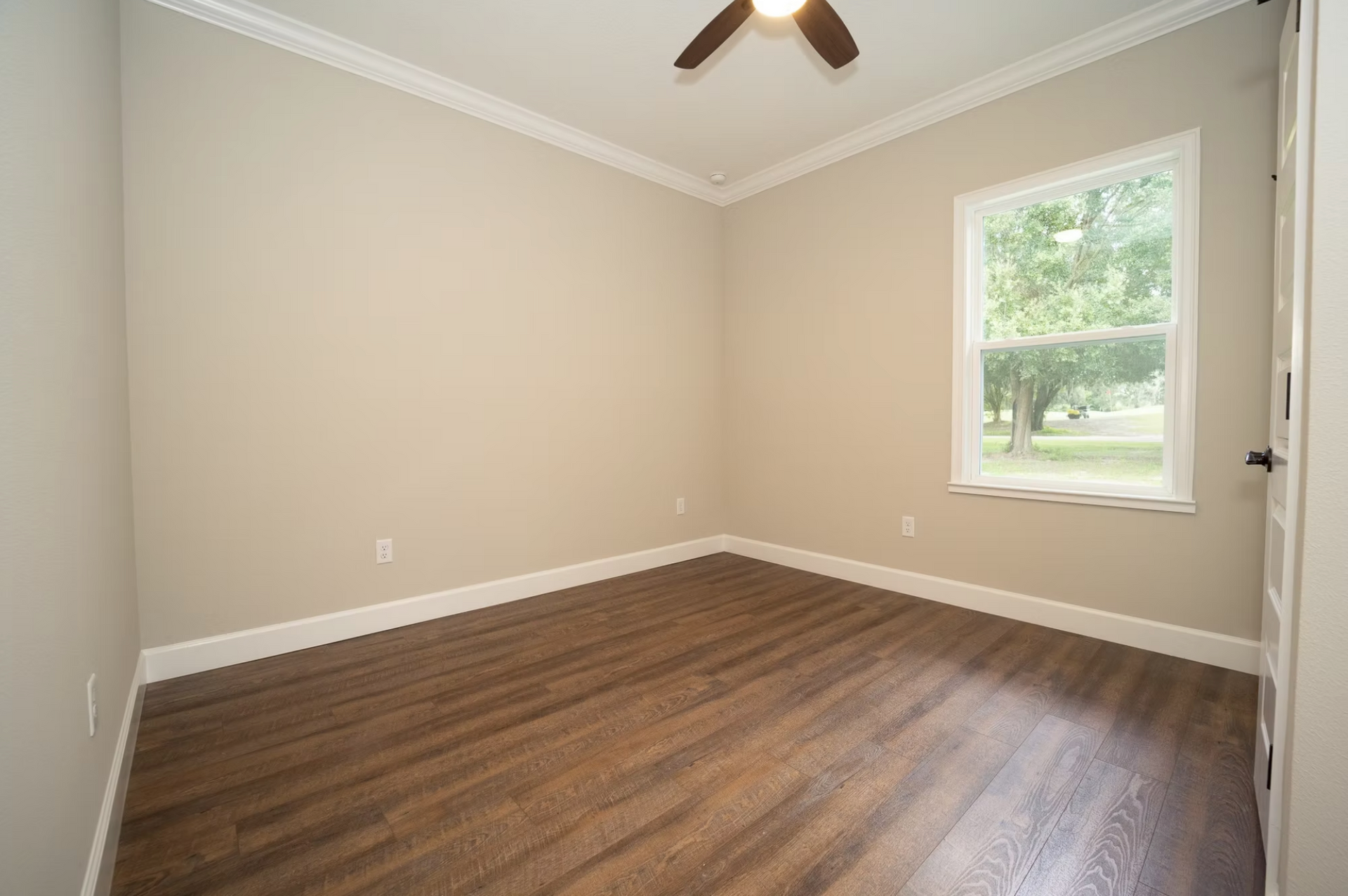 Empty beige bedroom with wood floors, ceiling fan, and a window overlooking greenery.