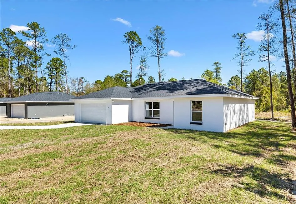 White single-story house with dark roof and attached garage, set on a grassy lot with trees and blue sky