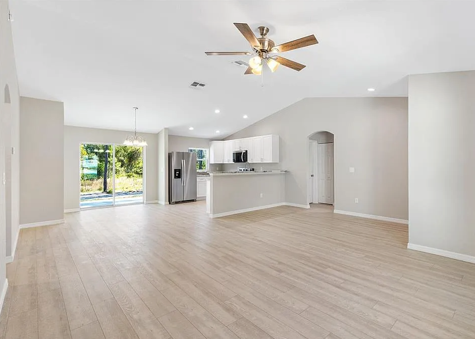 Empty open-concept living room with light wood floors, white kitchen, ceiling fan, and sliding glass door to patio