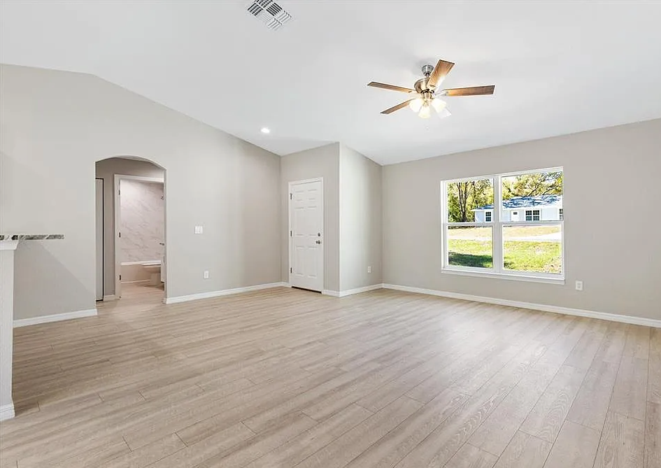 Empty beige living room with vaulted ceiling, ceiling fan, and large window letting in natural light