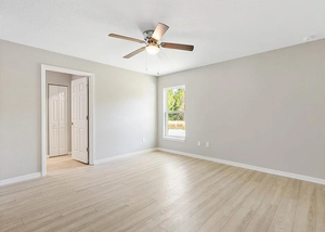 Empty light gray room with wood floors, ceiling fan, open doorway, and a narrow window.