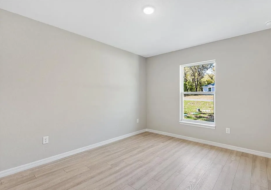 Empty beige bedroom with light wood floors and a single window overlooking greenery