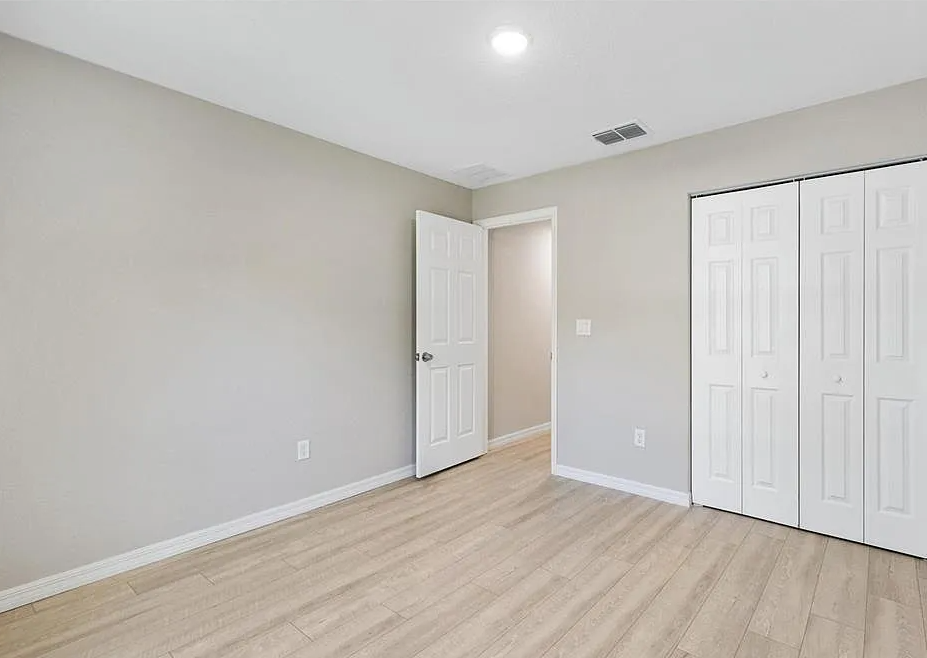 Empty beige bedroom with light wood floors, white closet doors, and an open door near the corner.