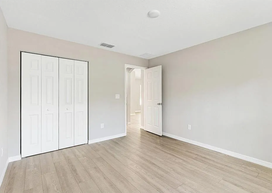 Empty beige bedroom with light wood floors, white closet doors, and an open doorway to a hallway
