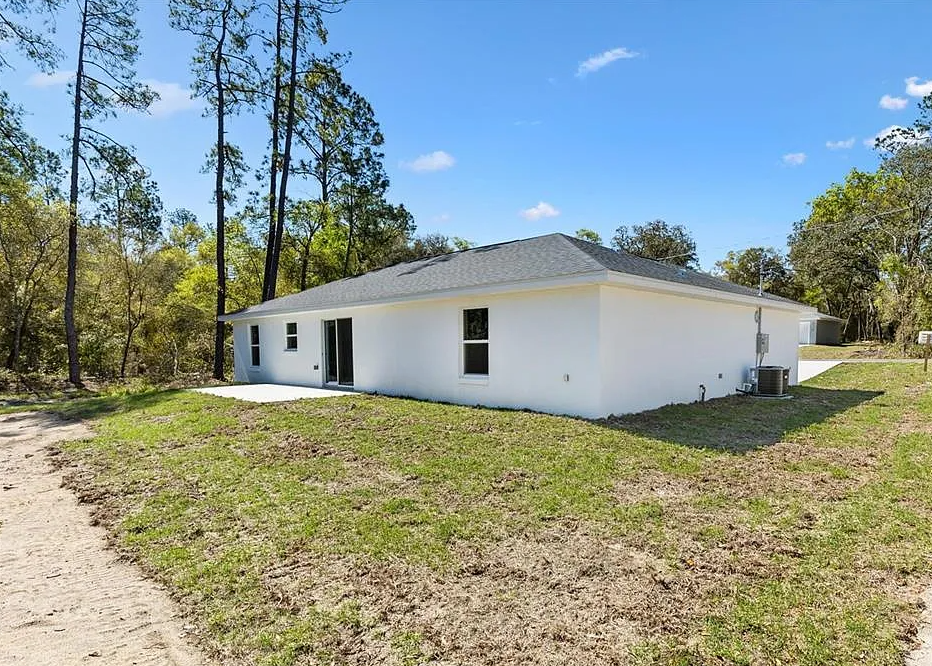 White single-story house with a gray roof in a grassy yard, surrounded by trees under a blue sky