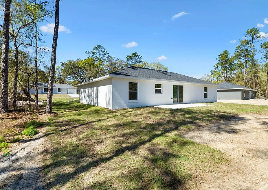 White single-story house with gray roof in a wooded yard under a clear blue sky