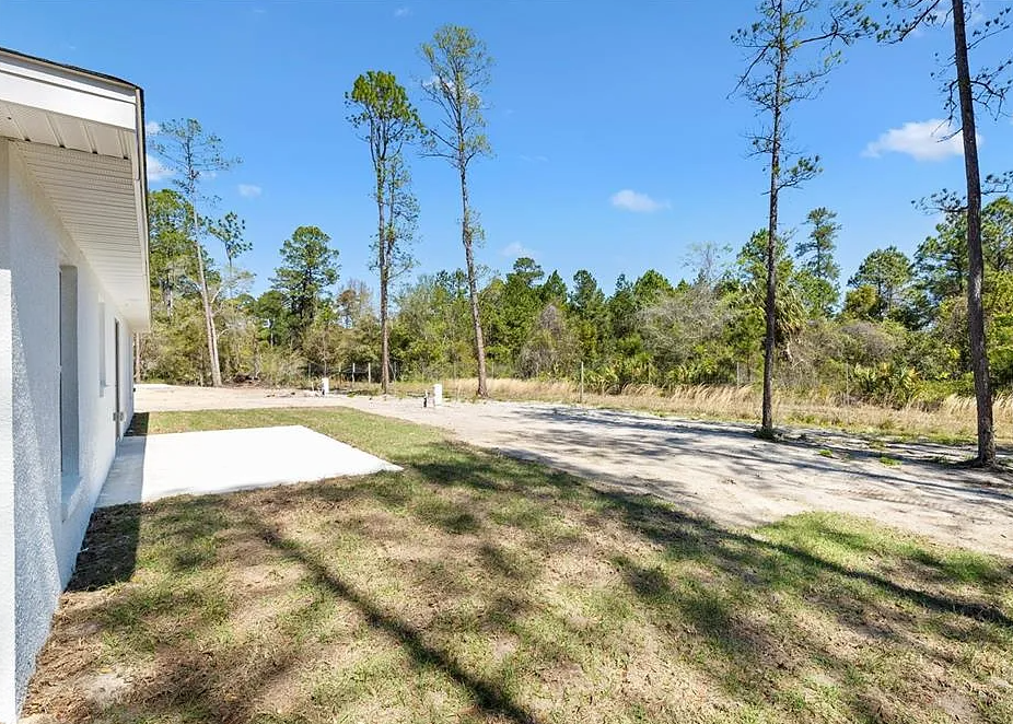 White house exterior with a concrete patio, grassy yard, and wooded landscape under a blue sky