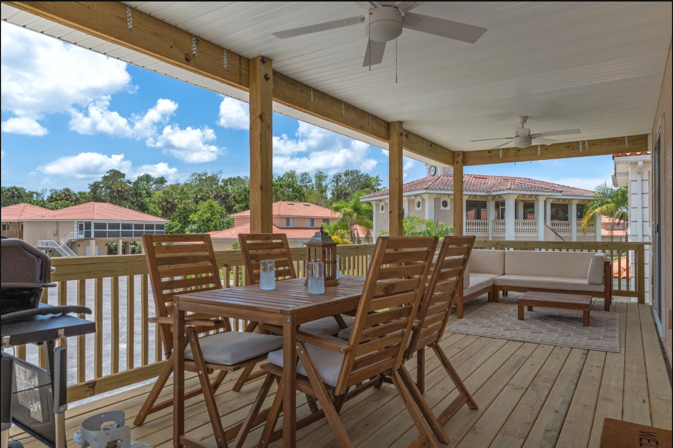 Covered wooden deck with dining table, chairs, and ceiling fan overlooking nearby homes and blue sky.