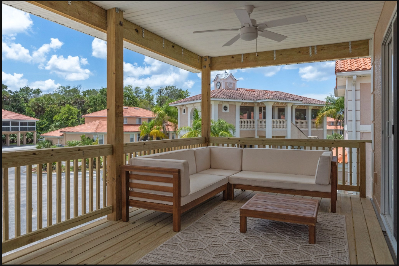 Covered wooden porch with beige sectional seating, coffee table, and view of nearby homes under a blue sky