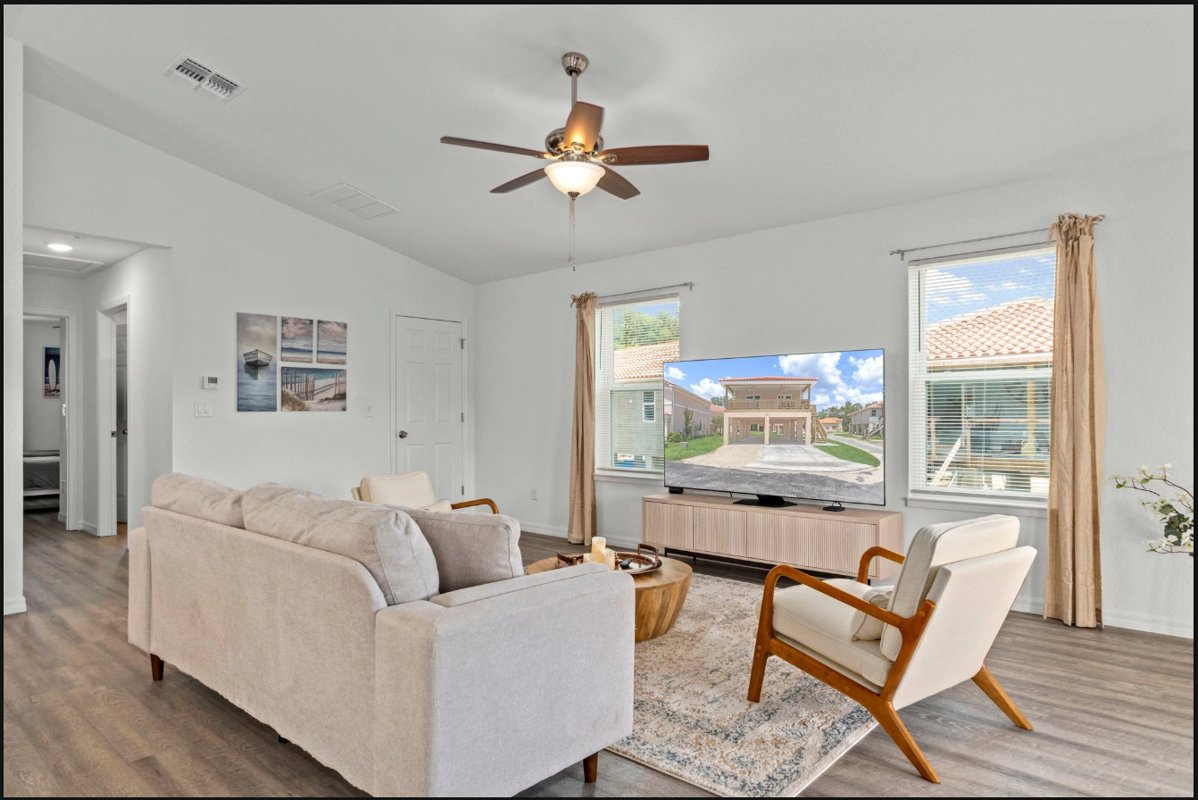 Bright living room with beige sectional, wood ceiling fan, and large windows overlooking a neighborhood.