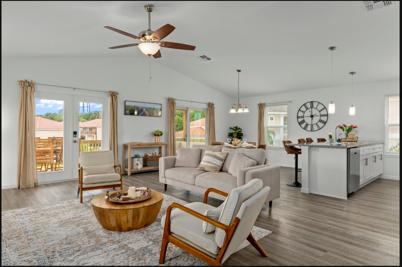 Bright open-plan living room and kitchen with beige furniture, wood floors, and large windows facing a mountain view.