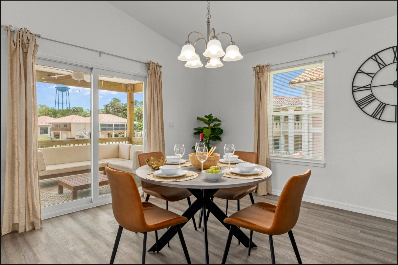 Bright dining room with round table, tan chairs, chandelier, large windows, and wall clock