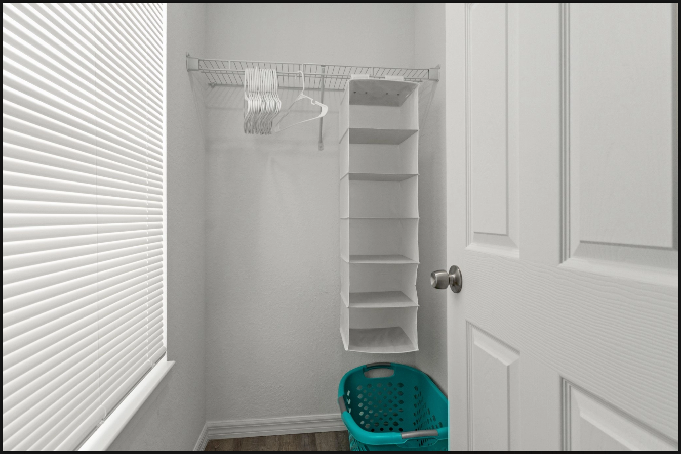 Small closet with wire shelves and hanging rods, partially open white door, and a teal laundry basket on the floor