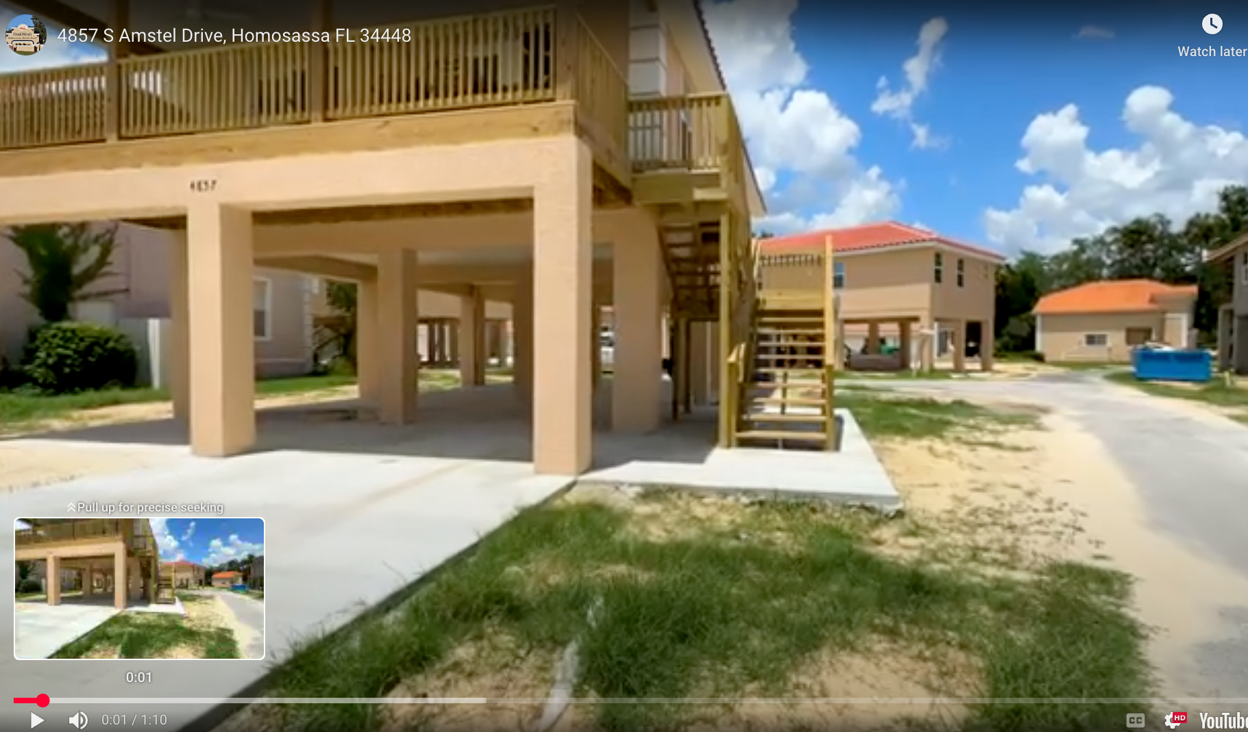 Stilted beach house with wooden deck, stairs, and sandy yard under a blue sky