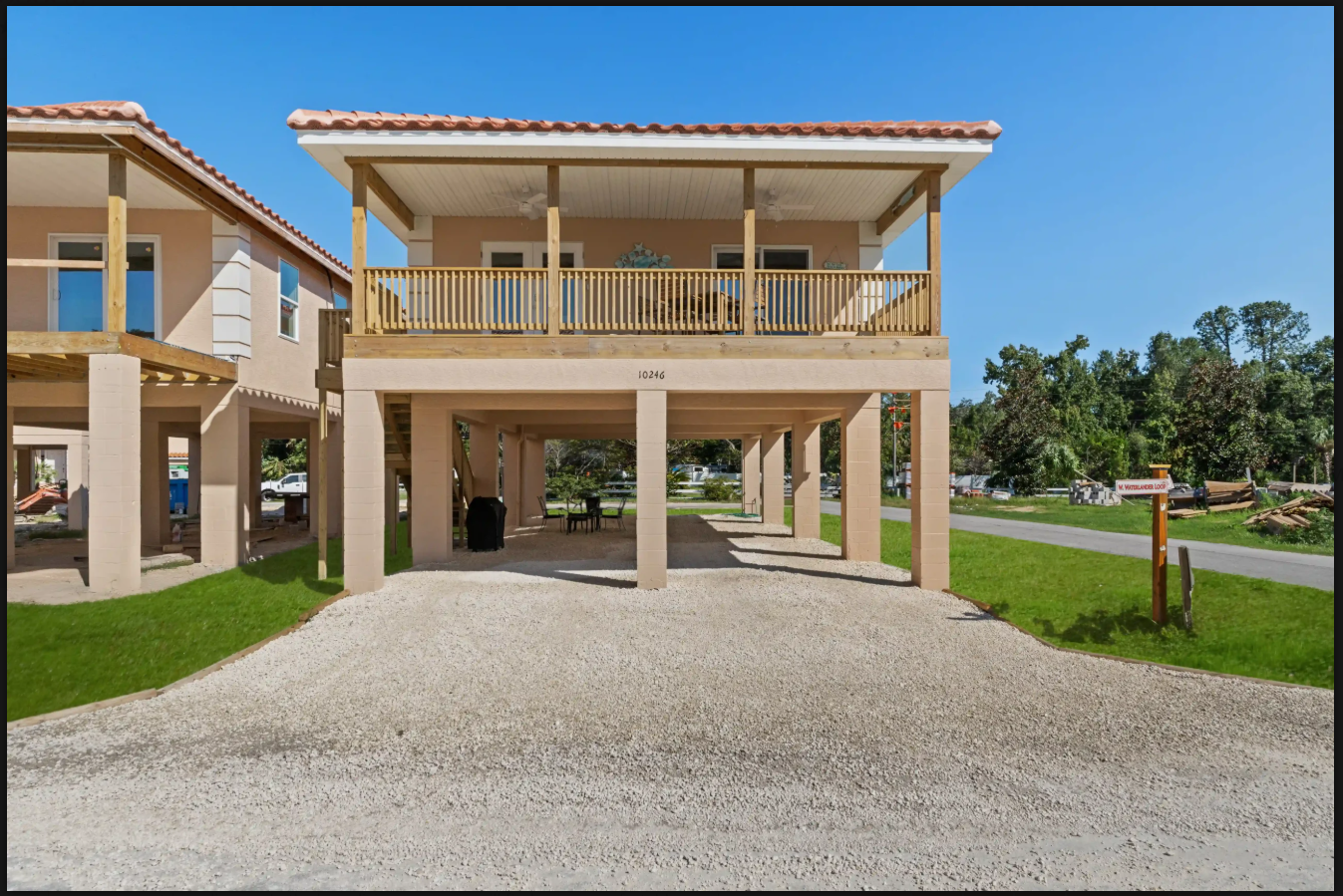 Raised beach house with beige columns, wraparound balcony, and gravel driveway under a blue sky