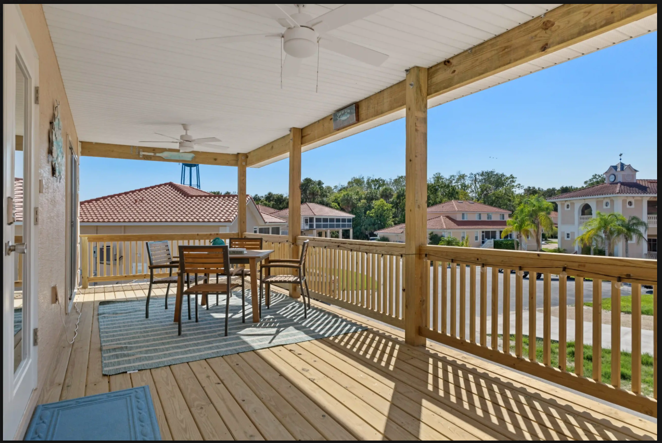 Covered porch with wooden railing, table, chairs, and ceiling fans overlooking suburban houses on a sunny day