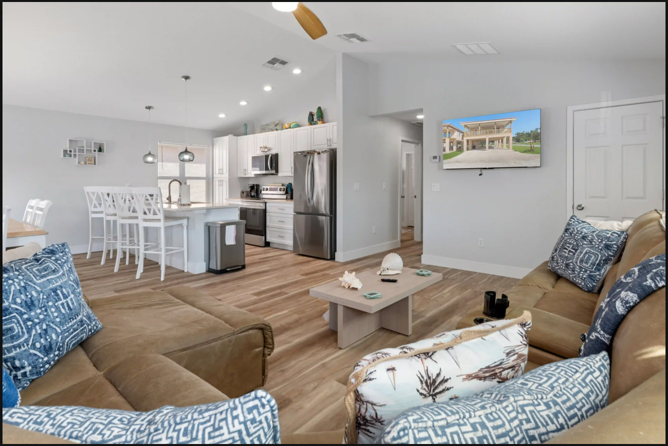 Bright open-concept living room and kitchen with tan sofas, blue patterned pillows, and wood floors