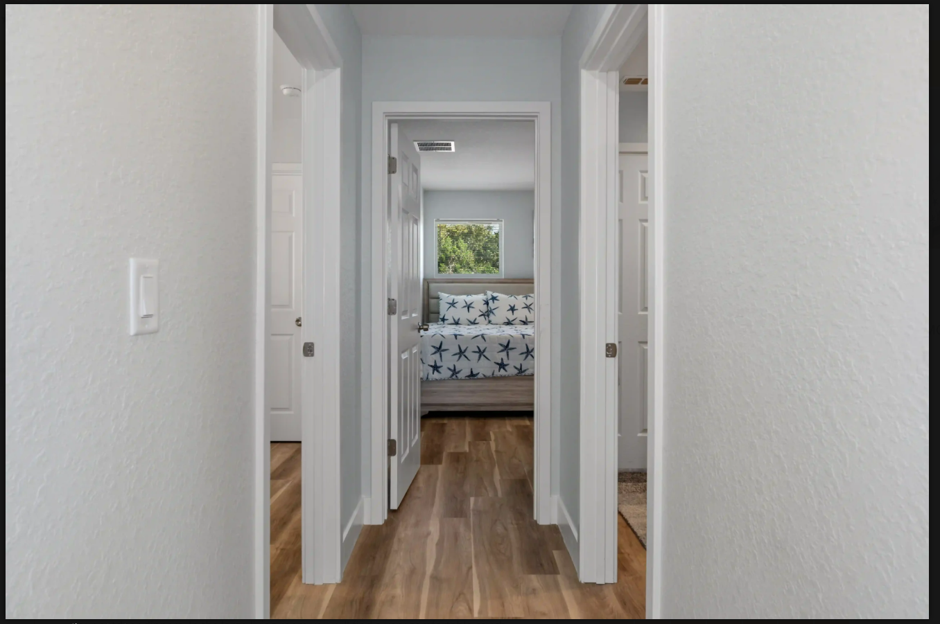 Hallway with white doors and wood floors, leading to a bedroom with a patterned bedspread and window.