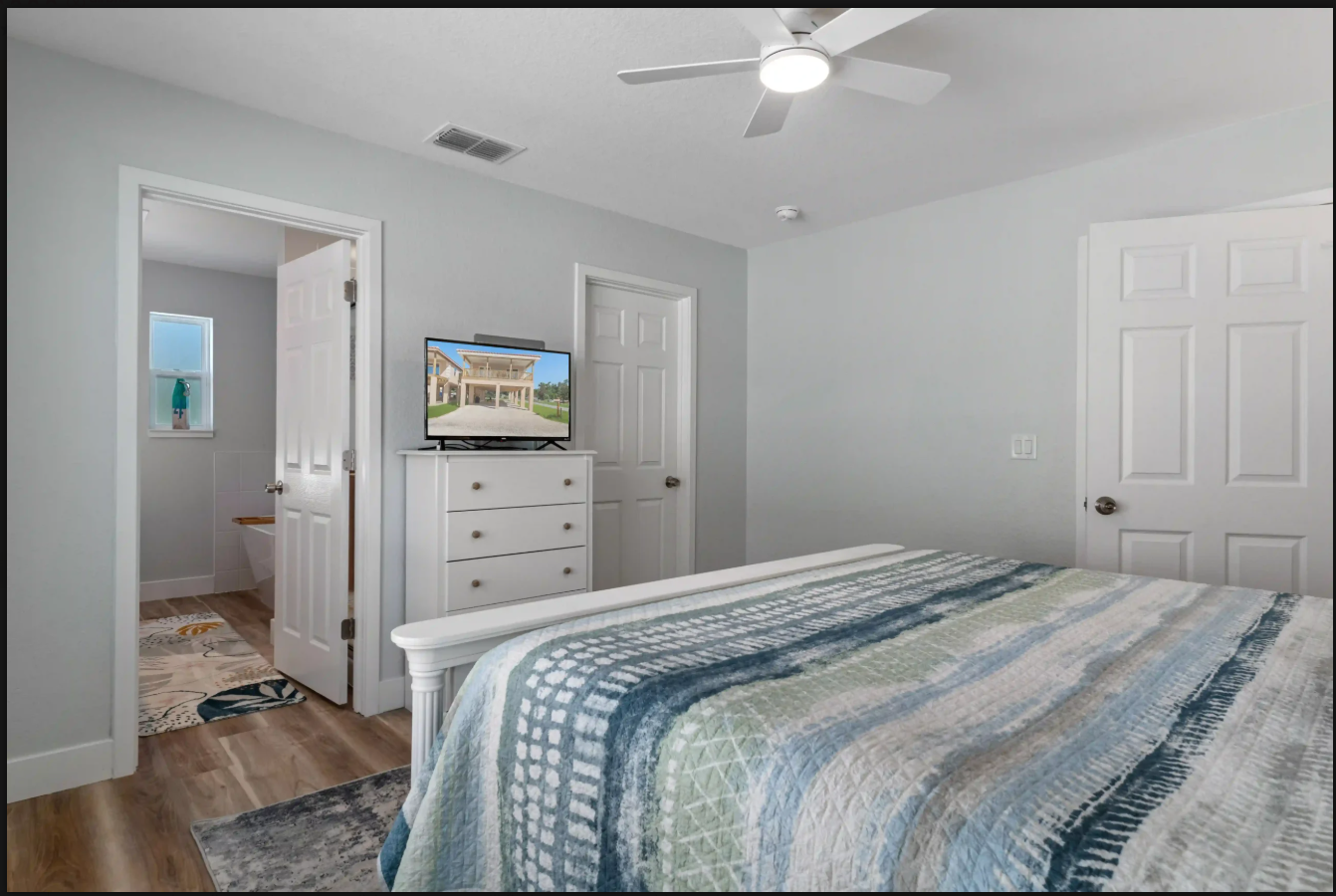 Bright bedroom with gray walls, blue patterned bedspread, white dresser, and open doorway to a bathroom