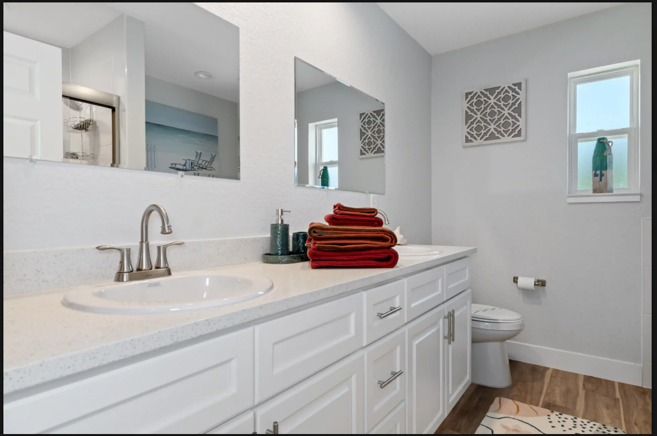 Bright white bathroom with double vanity, mirrors, stacked red towels, and a toilet near a window.