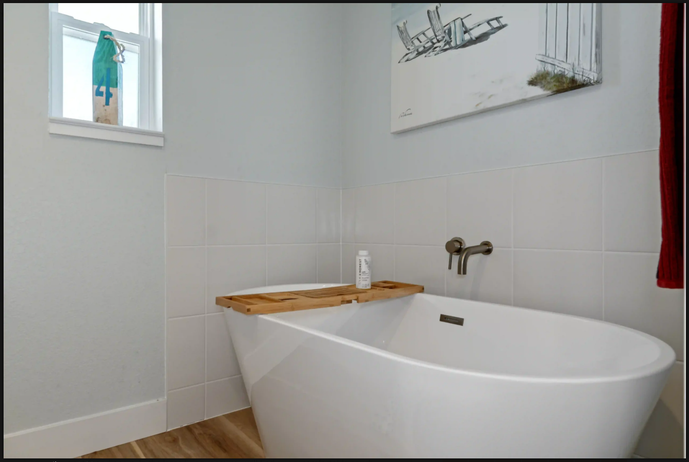 Modern bathroom with a freestanding white tub, wooden tray, wall art, and small window
