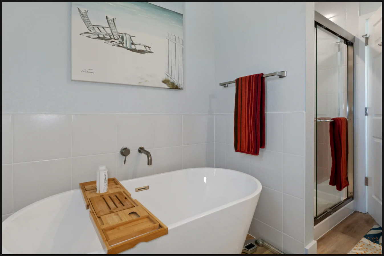 Modern bathroom with a white tub, wooden bath tray, red towels, and glass shower door