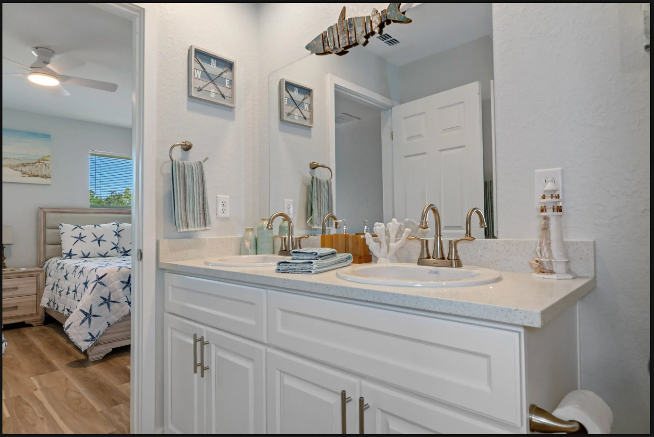 Bright bathroom vanity with white cabinets, double sinks, gold fixtures, and an open doorway to a bedroom.