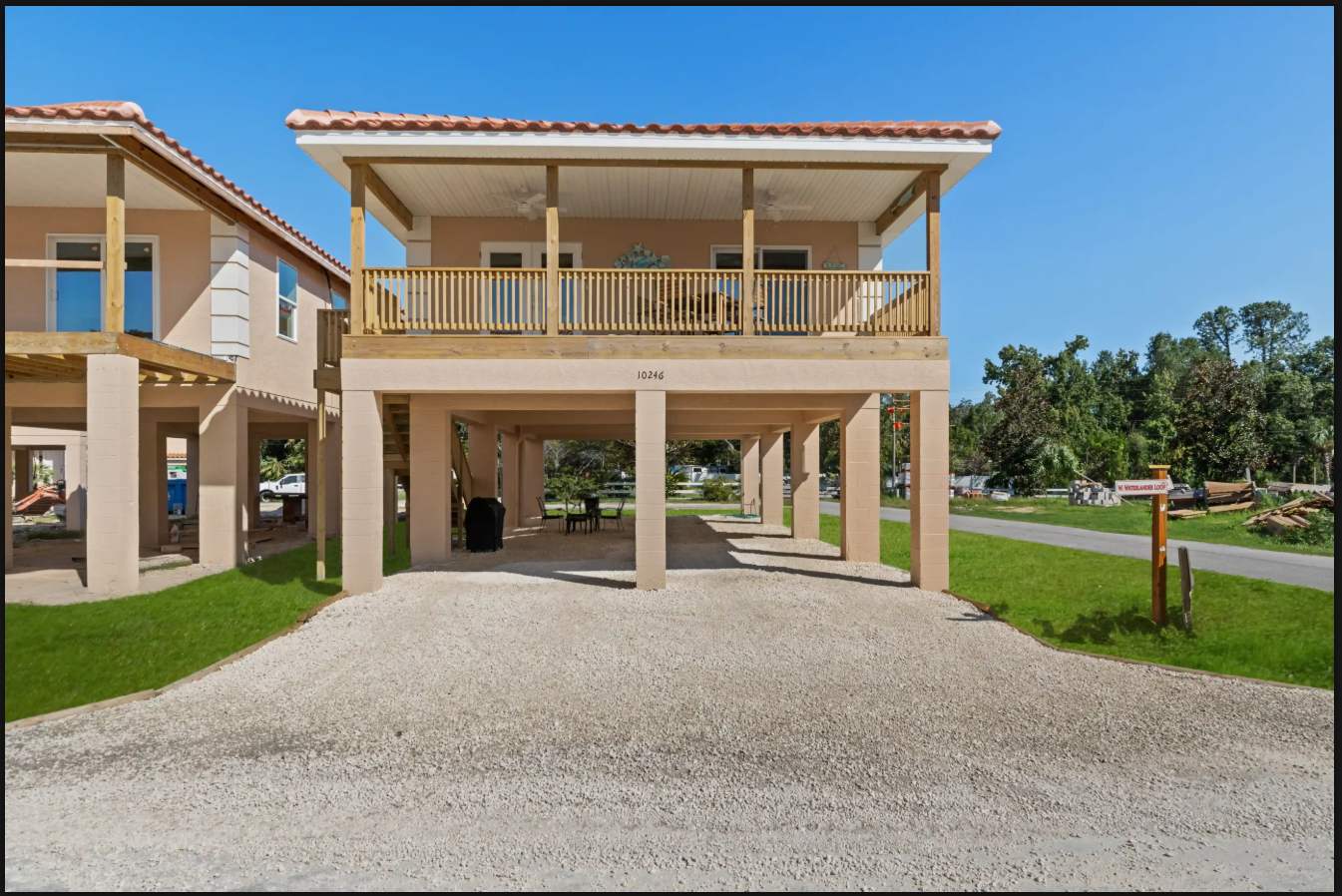 Two-story stilt house with a gravel driveway and open carport under a bright blue sky
