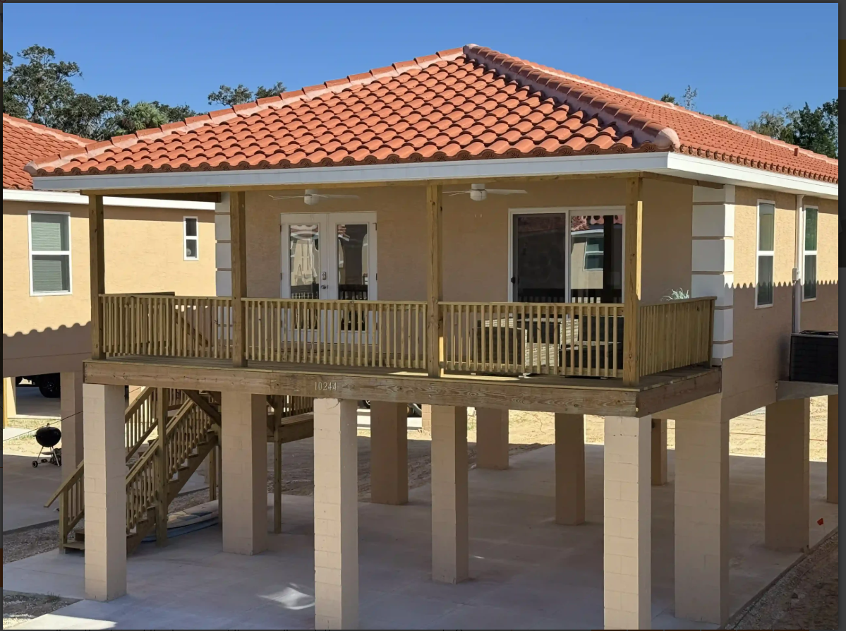 Elevated beach house with a red tile roof, wooden balcony, and stairs on stilts under a clear blue sky