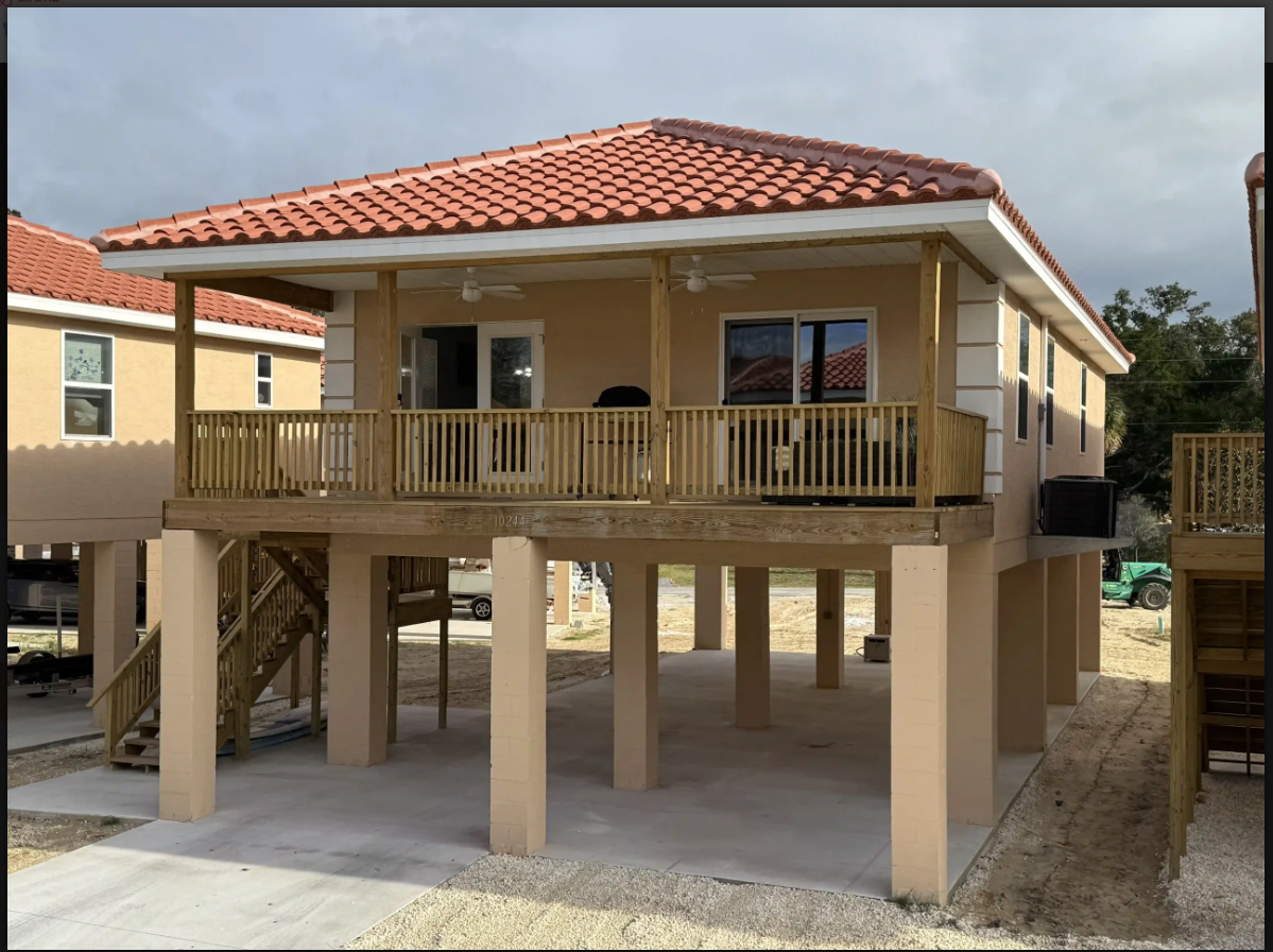 Elevated beige house with red tile roof and wraparound porch on stilts