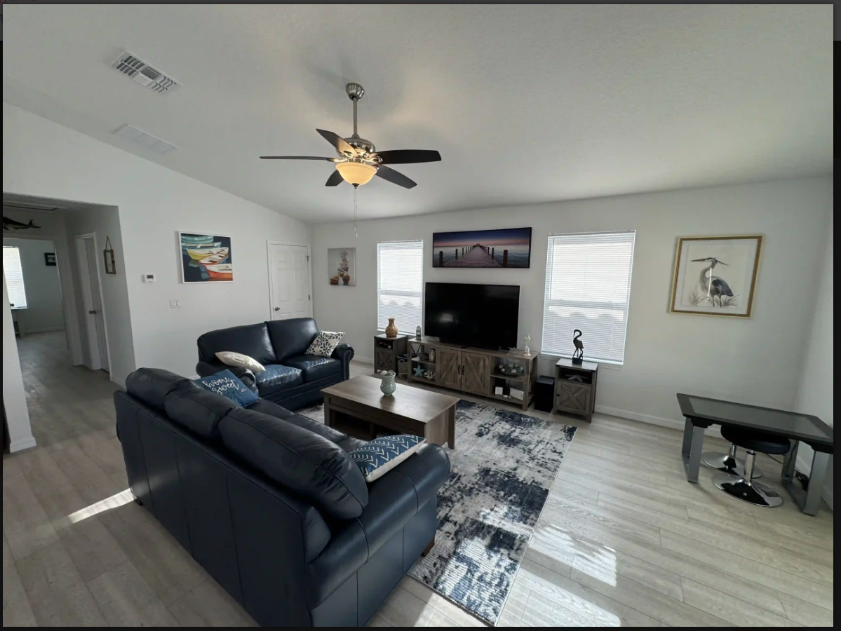 Bright living room with black sofas, TV, ceiling fan, wall art, and light wood floors