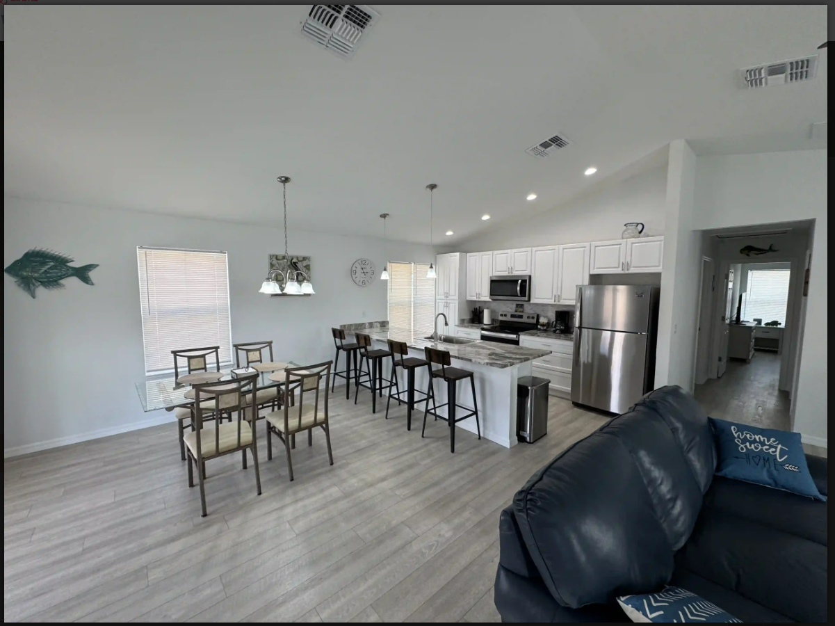 Open-concept kitchen and dining area with white cabinets, stainless fridge, bar stools, and a black sofa.