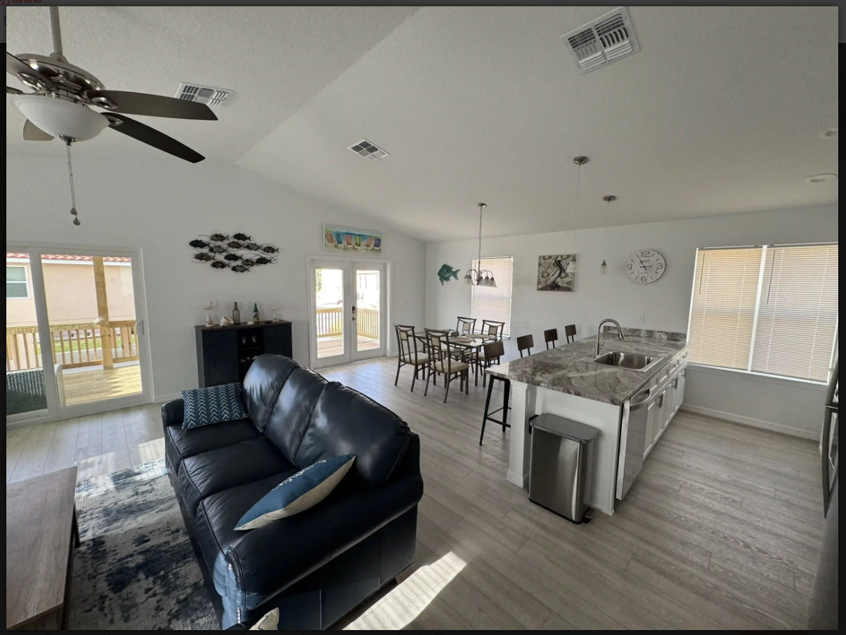Bright open-plan living room and dining area with black sofas, light wood floors, and large windows.