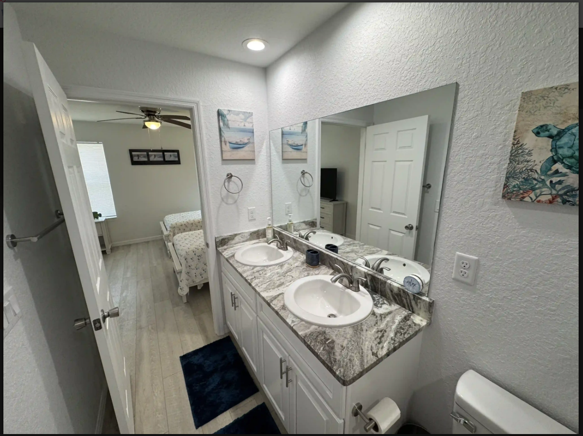 Bright bathroom with double vanity, large mirror, and doorway to a bedroom beyond.