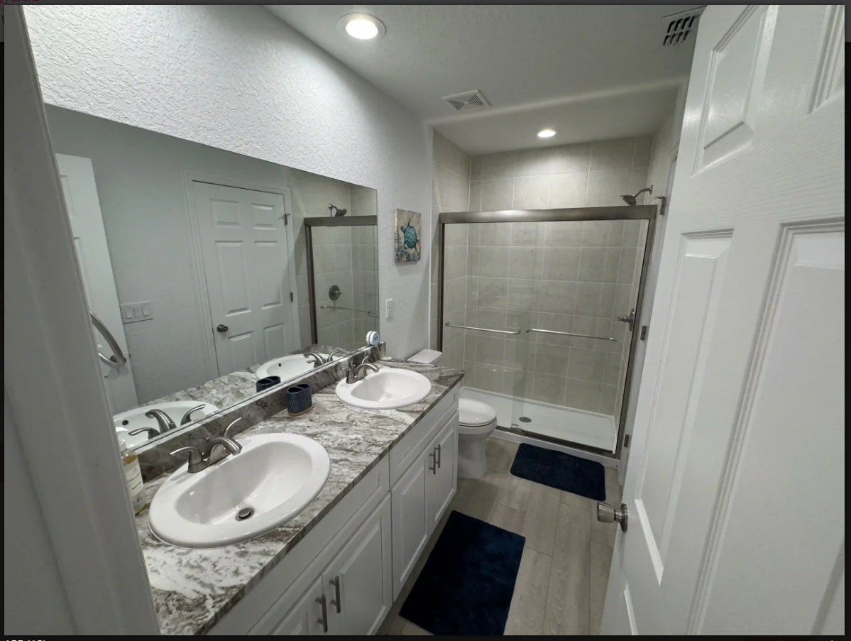Modern bathroom with double vanity, large mirror, glass shower, and dark floor mats