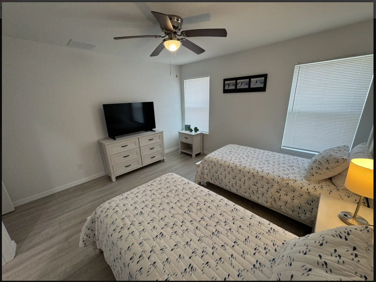 Bright bedroom with two beds, dresser, TV, ceiling fan, and windows with blinds