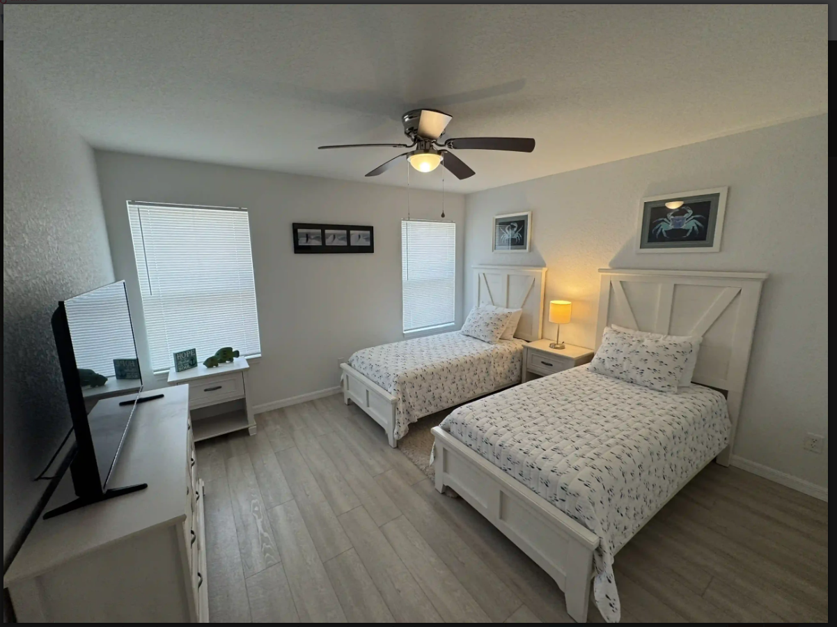 Bright twin bedroom with two white beds, nightstand lamp, ceiling fan, and windowed walls.