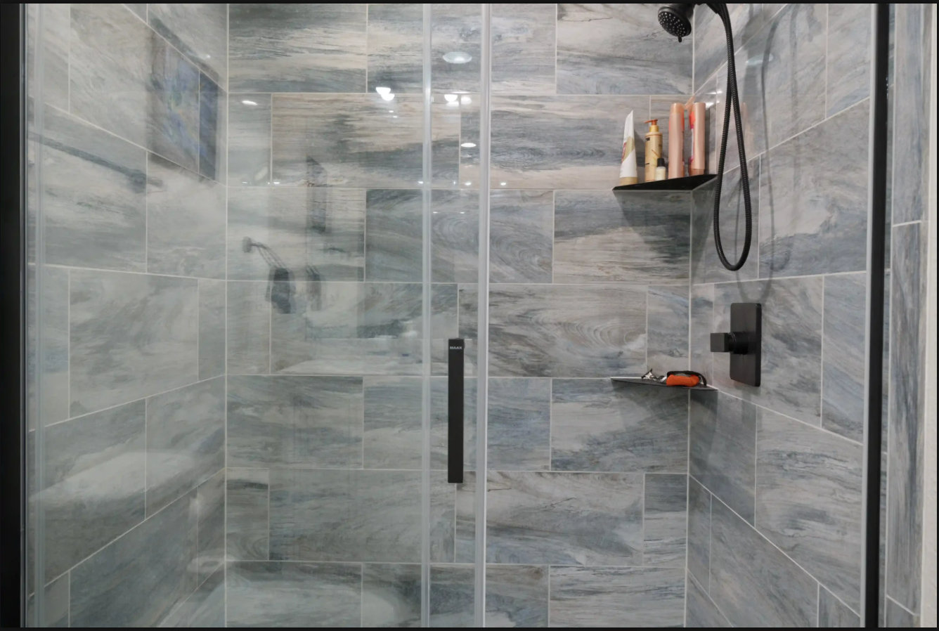 Glass shower with gray tile walls, corner shelf, and black shower fixtures