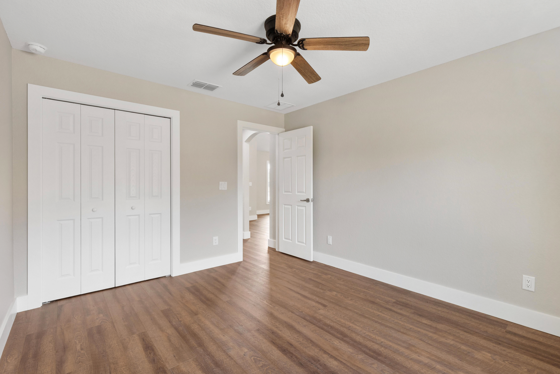 Empty beige bedroom with wood flooring, white closet doors, ceiling fan, and open doorway to hall