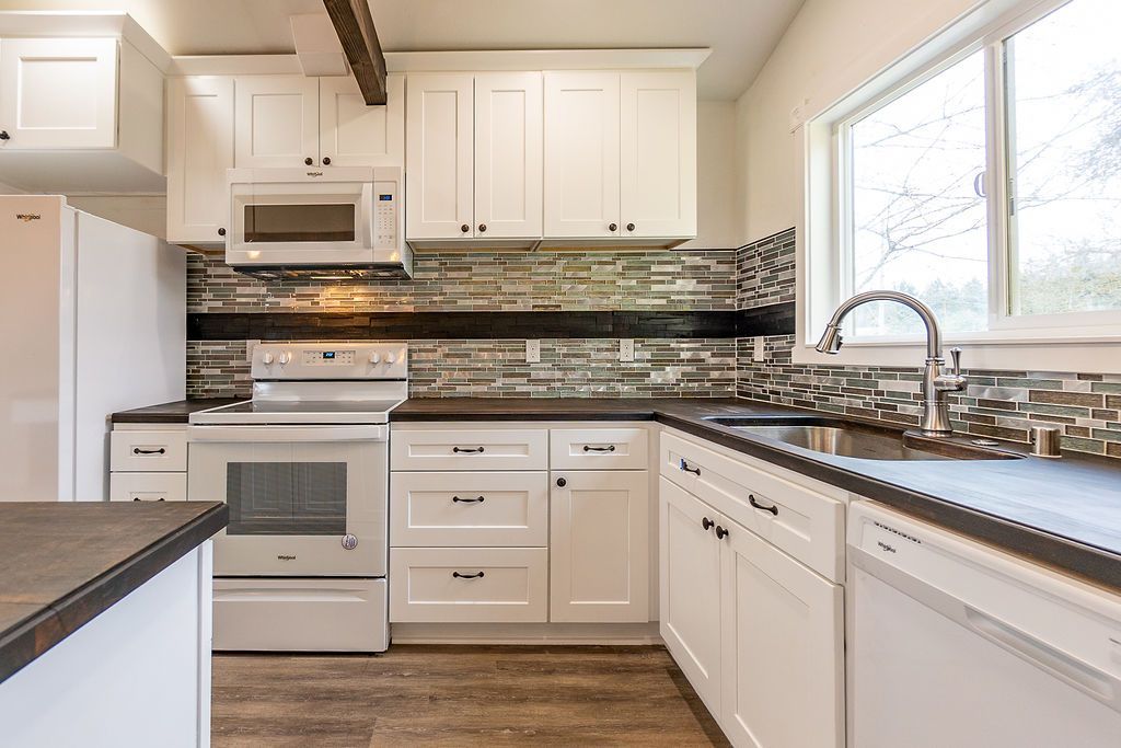 White kitchen with cabinets, stove, microwave, and sink; stone backsplash, dark countertops, and a window.