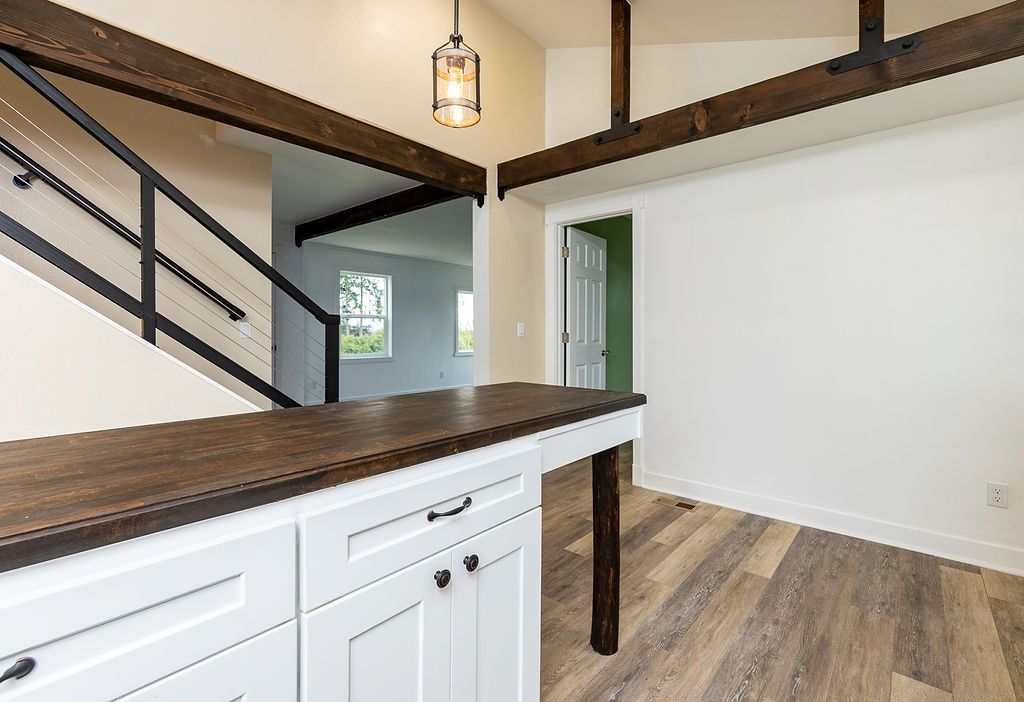 Kitchen with white cabinets, dark wood countertop and beams. A staircase with black railing is visible.