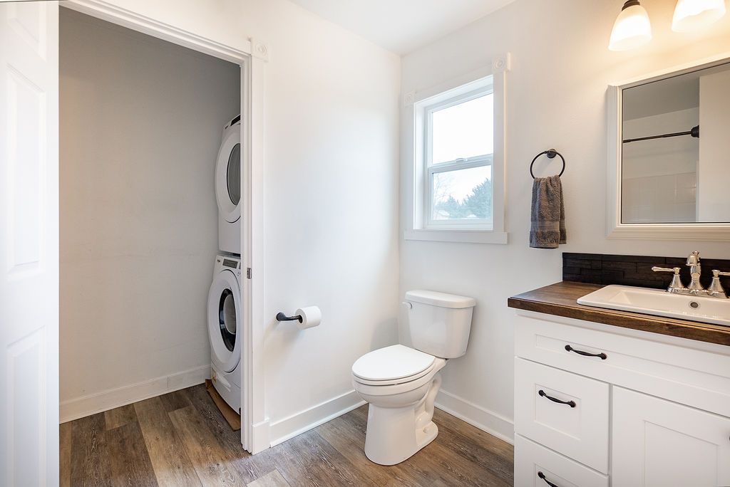 Bathroom with stacked washer/dryer, toilet, sink, and window. White walls, wood floor, and vanity.