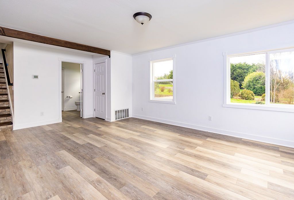 Empty living room with wood-look flooring, white walls, and a view of trees through windows.